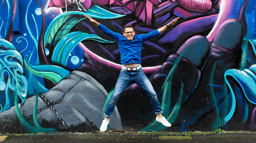 Nev Harris Jumping In Front Of A Blue and Purple Wall Of Graffiti
