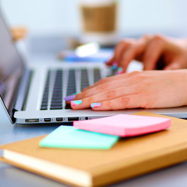 Freelancing female typing on laptop with a notebook and post-its in the foreground to represent increased productivity with support and accountability through Slack